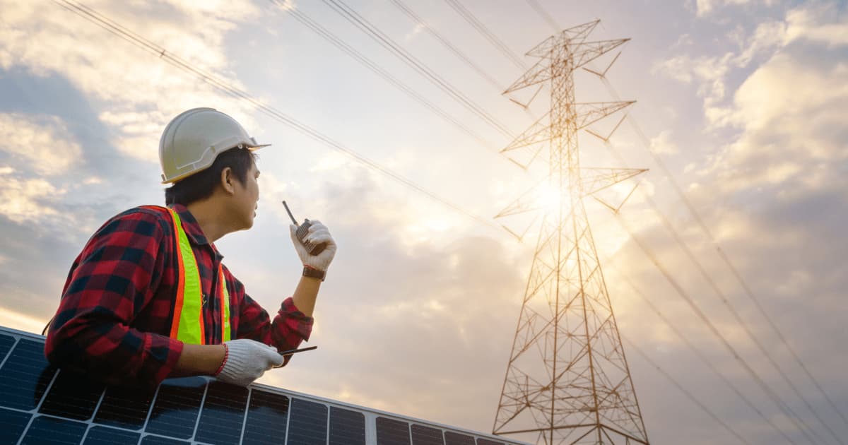 Person mit Walkie-Talkie in Arbeitskleidung und Bauhelm lehnt an PV-Modulen und schaut zu einem Strommasten hinauf. Im Gegenlicht der Sonne.